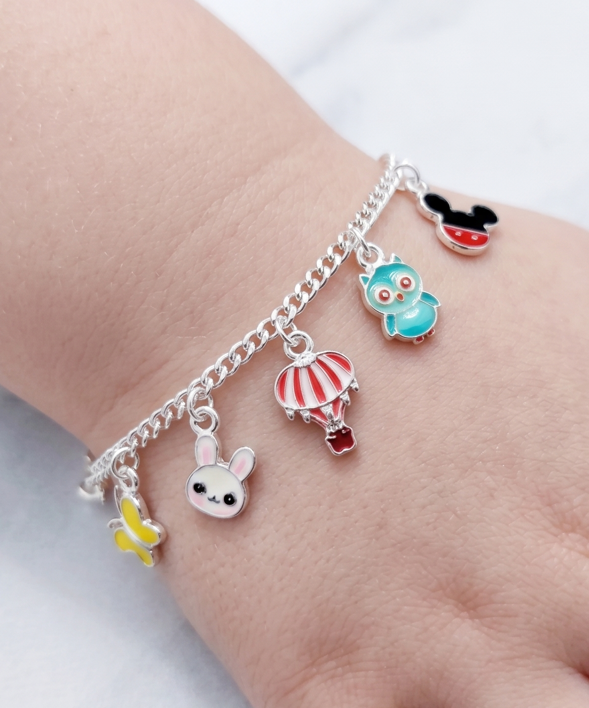 A silver chain bracelet with five colorful cartoon charms, including a hot air balloon, owl, mouse, bunny, and butterfly, shown worn on a child's wrist against a light marble background.