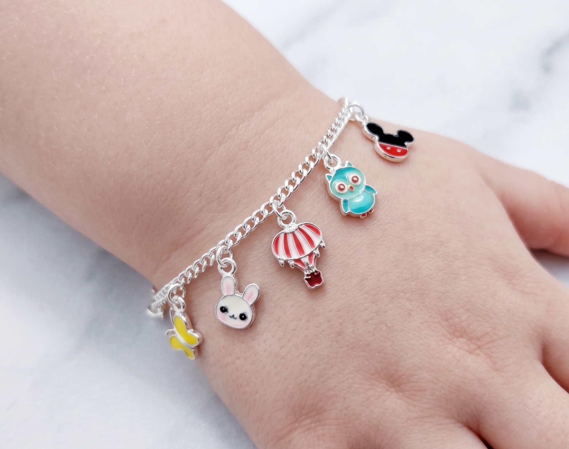 A silver chain bracelet with five colorful cartoon charms, including a hot air balloon, owl, mouse, bunny, and butterfly, shown worn on a child's wrist against a light marble background.