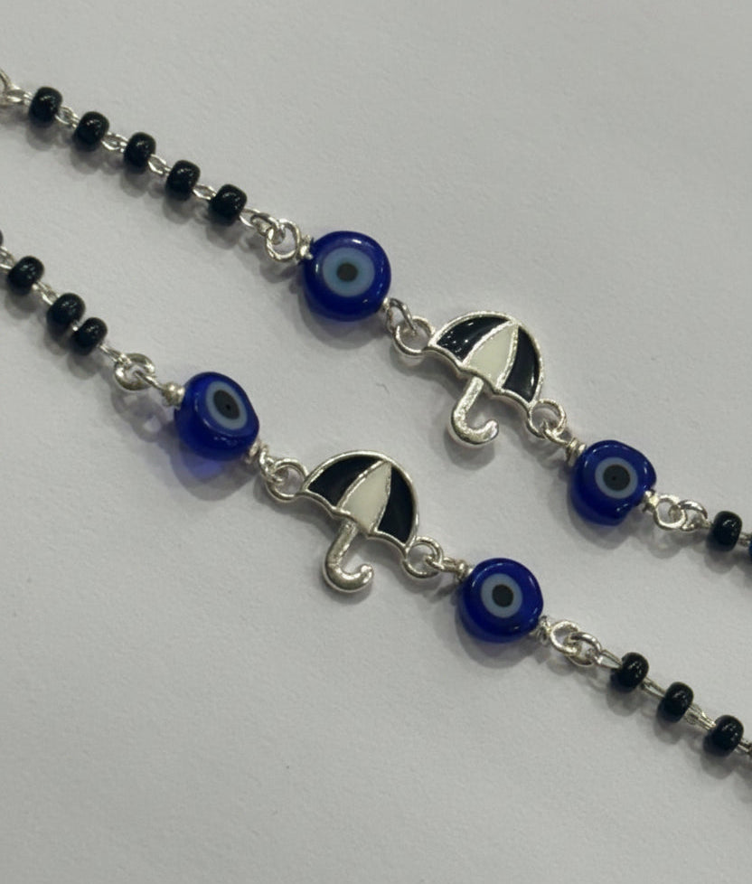 A close-up of a silver beaded necklace featuring evil eye glass beads and silver umbrella charms, laid out on a light gray background.
